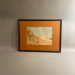 May include: Framed watercolor painting of a desert landscape. The artwork features a sandy foreground, a mesa, and a blue sky with a few birds. The painting is set within an orange mat and a black frame.
