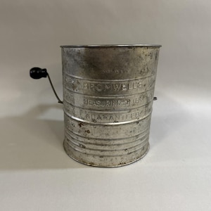 May include: Vintage metal sifter with a black handle. The sifter has the words "BROMWELL'S MEASURING SIFTER GUARANTEED" embossed on the side. The sifter has measurements for cups. A classic kitchen tool.