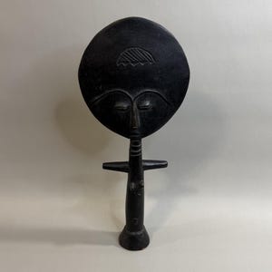 May include: A dark brown, hand-carved wooden figurine featuring a large, circular head with stylized facial features, including arched eyebrows and closed eyes. The figure has a slender, cylindrical body with horizontal neck rings and arms.