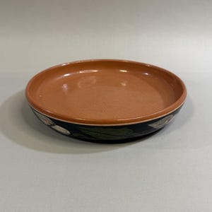 May include: A terracotta-colored ceramic bowl with a black rim featuring a floral design. The bowl has a shallow, round shape, suitable for serving or display. The interior is a warm, earthy tone.