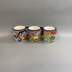 May include: Three ceramic mugs with colorful floral designs. Each mug features a different pattern with a blue rim and handle. The mugs have a white interior and are set against a gray background.