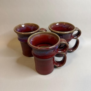 May include: Three ceramic mugs with a glossy deep red glaze and subtle blue-toned highlights. Each mug features a cylindrical body, a flared rim, and a curved handle. The rims and handles are accented with a brownish-tan finish, creating a contrast.