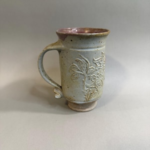 May include: A tall ceramic mug with a speckled light gray glaze and a pink interior. The exterior features a raised, embossed floral pattern on the side. It has a curved handle and a flared rim, with a distinct footed base at the bottom.