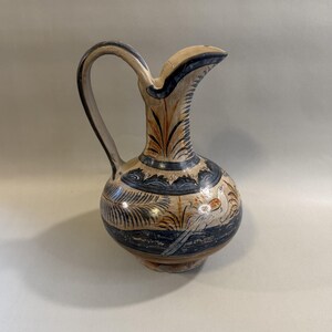 May include: A ceramic pitcher with a curved handle and spout. The pitcher is painted with a beige, blue, and brown design featuring stylized leaves and birds. The base is a darker brown color.