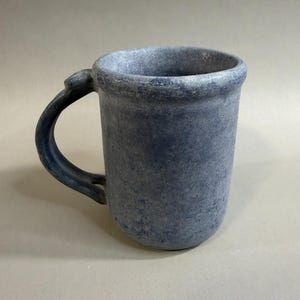 May include: A blue ceramic mug with a textured surface and a curved handle. The mug has a cylindrical shape and a slightly flared rim. The handle is attached to the side of the mug and is also blue.