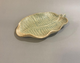 May include: A handmade ceramic leaf-shaped dish in a pale green color. The dish has a textured surface with detailed veining and a slightly raised edge. The leaf dish is a decorative item.