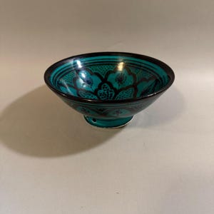 May include: A small, circular ceramic bowl featuring a vibrant turquoise glaze with intricate, dark black floral and geometric patterns painted across the interior and exterior surfaces. The bowl rests on a raised, narrow circular base.