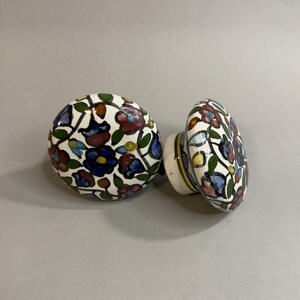 May include: Two round ceramic knobs featuring a hand-painted floral pattern with dark blue, maroon, and green petals on a white background. Each knob has a smooth, glossy finish and a short cylindrical base for mounting to furniture or cabinetry.
