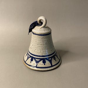 May include: A ceramic bell with a speckled, off-white finish and blue painted details. The bell has a loop at the top and a dark blue ribbon. The base features a repeating blue pattern. A decorative item.