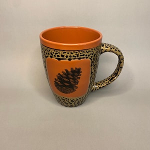 May include: A ceramic mug with an orange interior and a decorative exterior. The exterior features a black and gold spotted pattern and an orange square with a pinecone illustration. The handle is also decorated with the spotted pattern.