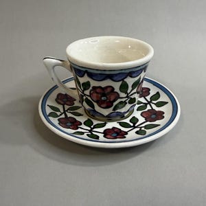 May include: A ceramic teacup and saucer set featuring a hand-painted floral pattern with dark red flowers, green leaves, and blue scalloped borders on a cream-colored background. The cup has a curved handle and the saucer has a matching floral design.