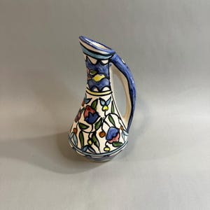 May include: A ceramic pitcher with a cream-colored base featuring hand-painted floral motifs in blue, red, yellow, and green. The vessel has a tapered neck, a curved handle, and a flared spout, decorated with horizontal bands of blue and black lines.