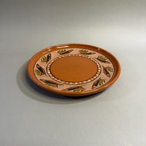 May include: A round, terracotta-colored ceramic plate with a central oval and a patterned border. The border features a repeating design of stylized fish in blue, green, and white. The plate has a slightly raised rim.