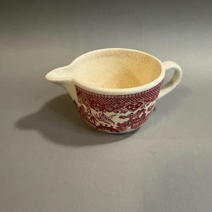 May include: Cream-colored creamer with a red Willow pattern design. The creamer has a spout and a handle. The interior has a cracked appearance. The red pattern wraps around the exterior of the creamer.