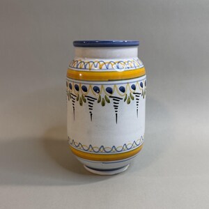 May include: A cylindrical ceramic vase with a white base, featuring hand-painted horizontal bands in yellow and blue. The design includes decorative patterns of blue dots, green leaf-like motifs, and thin black vertical lines around the body.