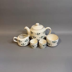 May include: A cream-colored ceramic tea set featuring a repeating dark green shamrock pattern and thin gold trim. The set includes a teapot with a lid, a creamer pitcher, two small cups, and a sugar bowl, all arranged together on a flat surface.