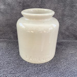 May include: A white ceramic jar with a rounded body and a slightly flared top. The jar has a raised inscription that reads "LAB-Lagny".