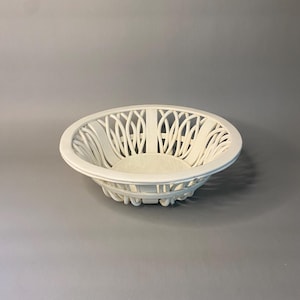 May include: A white, oval-shaped basket with a decorative openwork design. The basket has a rounded rim and a solid base. The openwork pattern features vertical and curved lines, creating an elegant and airy appearance. The basket is made of a solid material.