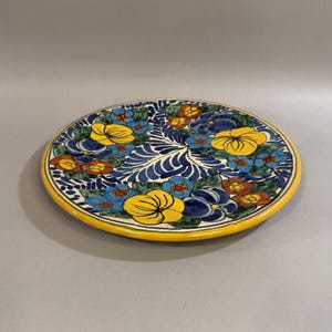 May include: A round, hand-painted ceramic plate with a yellow rim. The plate features a floral design with yellow, blue, and orange flowers on a white and blue background. The plate is a decorative item.