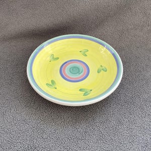 May include: A yellow ceramic bowl with a blue and green rim and a multi-colored circle design in the center. The bowl has a green leaf pattern around the rim.