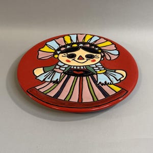 May include: A round ceramic plate with a bright red background featuring a central illustration of a doll with black hair, braided pigtails, and a colorful dress with pink, green, and yellow stripes, accented by a small red heart on the bodice.