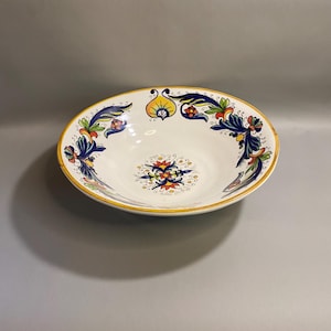 May include: A white ceramic serving bowl with a yellow rim and intricate floral designs in blue, yellow, and red. The bowl has an oval shape and is decorated with symmetrical patterns.