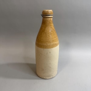 May include: A vintage stoneware bottle featuring a two-tone design with a glossy tan glaze on the upper neck and shoulder, transitioning to a matte off-white finish on the cylindrical body. A thin metal wire is wrapped around the bottle neck.