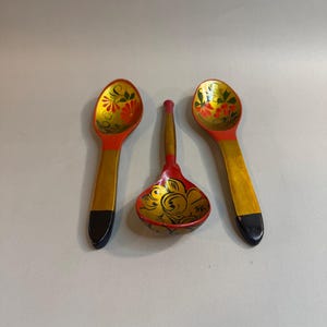 May include: Three wooden spoons featuring hand-painted floral motifs in red, green, and gold. Two spoons are elongated with black handles, while the central spoon is shorter and rounder with a gold handle, all displaying intricate decorative patterns.