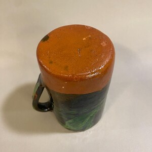 Puede incluir: Una taza de cer&aacute;mica hecha a mano con una parte superior de color naranja oscuro y una base negra. La base presenta dise&ntilde;os de hojas verdes. La taza tiene un asa negra y probablemente se usa para beber bebidas.