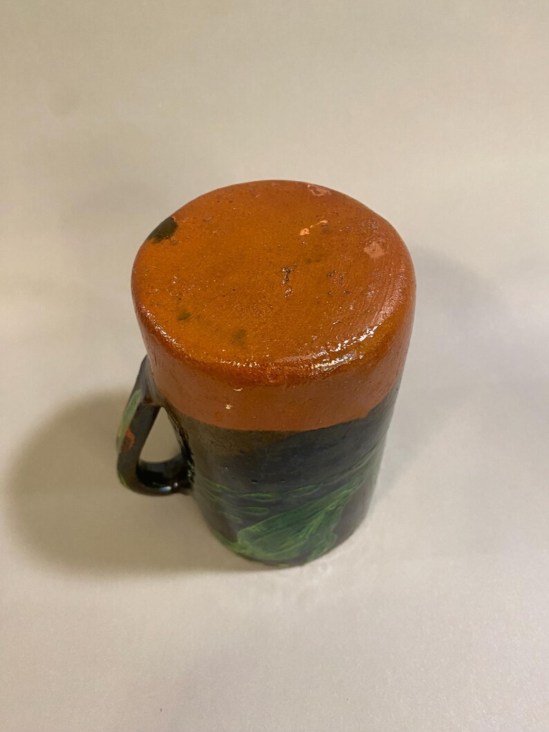 Puede incluir: Una taza de cer&aacute;mica hecha a mano con una parte superior de color naranja oscuro y una base negra. La base presenta dise&ntilde;os de hojas verdes. La taza tiene un asa negra y probablemente se usa para beber bebidas.