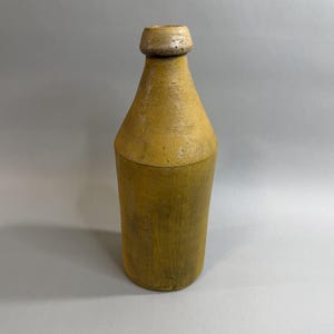May include: A tall, cylindrical stoneware bottle with a mustard yellow finish and a tapered neck. The bottle features a thick, rounded rim at the top opening. The surface has a matte, textured appearance with subtle variations in the yellow tone.