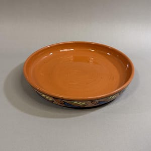 May include: A round, terracotta-colored ceramic plate with a wide rim. The rim features a decorative border with blue, green, and brown patterns. The plate has a smooth, glossy surface.
