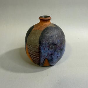 May include: A spherical ceramic vase with a narrow neck and a textured surface. The exterior features a blend of earthy orange, deep black, and mottled blue glazes, with a section displaying a series of horizontal incised lines around the body.