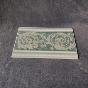 May include: A rectangular decorative tile with a white border and a green and white floral design. The tile has a textured surface and is likely used for interior design or home decor.