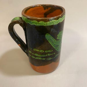 Puede incluir: Taza de cer&aacute;mica pintada a mano con exterior marr&oacute;n oscuro, interior naranja y detalles verdes. La taza tiene un asa y un dise&ntilde;o verde decorativo alrededor del borde y en el lateral. La base es naranja.