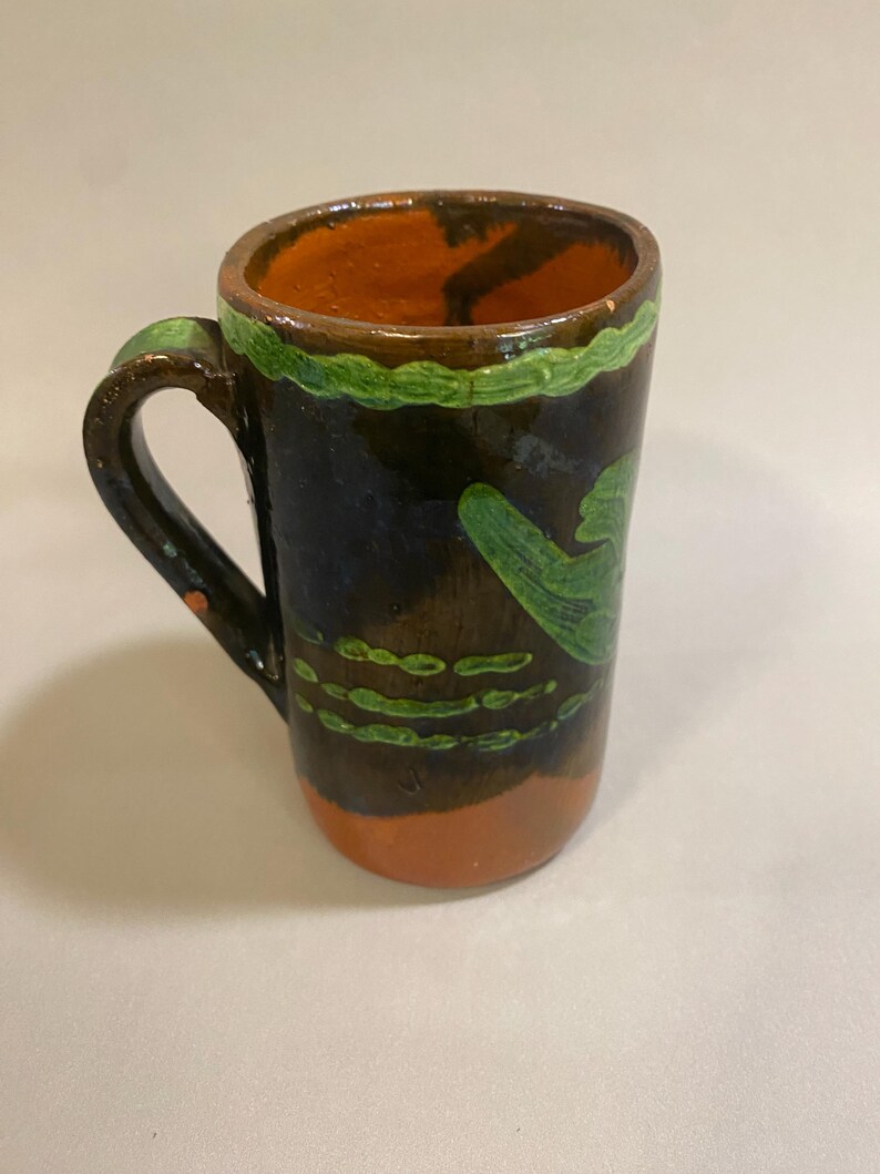 Puede incluir: Taza de cer&aacute;mica pintada a mano con exterior marr&oacute;n oscuro, interior naranja y detalles verdes. La taza tiene un asa y un dise&ntilde;o verde decorativo alrededor del borde y en el lateral. La base es naranja.