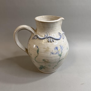 May include: A ceramic pitcher with a cream-colored glaze, featuring a curved handle and a pouring spout. The body is decorated with hand-painted blue floral motifs and green vines, accented by a raised, sculpted blue wave-like trim around the neck.