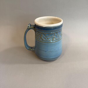 May include: A handmade ceramic mug with a light blue glaze. The mug features a decorative leaf pattern around the top and a cream-colored interior. The handle has a small decorative element. A unique piece for enjoying beverages.