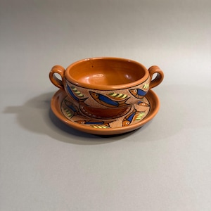 May include: A terracotta soup bowl with two handles and a matching saucer. The bowl and saucer are decorated with a repeating pattern of stylized fish in blue, yellow, and green. The set is likely handmade and has a rustic aesthetic.