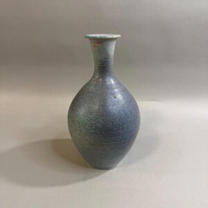 May include: A ceramic vase with a bulbous body and a narrow neck. The surface features a mottled, textured finish in shades of dark blue, grey, and hints of green. The rim is flared and displays a subtle light pinkish hue along the top edge.