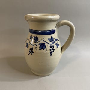 May include: A cream-colored ceramic pitcher featuring a hand-painted dark blue horizontal band around the neck and a decorative vine motif with leaves and grape clusters encircling the body, complete with a curved handle and a pouring spout.