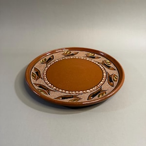 May include: A round, brown ceramic plate with a decorative border. The border features a repeating leaf design in shades of brown, green, and black. The center of the plate is a solid brown color. The plate is likely handmade.