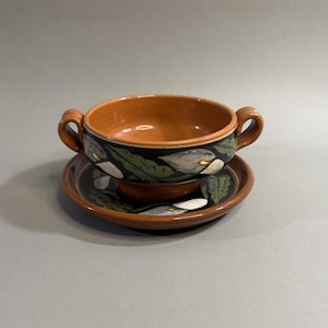 May include: A ceramic soup bowl with two handles and a matching saucer. The bowl and saucer are brown with a black band featuring white calla lilies and green leaves. The interior of the bowl is a lighter brown.