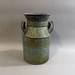 May include: A vintage-style metal milk can with a distressed finish. The can features a cylindrical body with horizontal bands, two arched handles, and a flared top. The color is a mix of green, gold, and black, giving it a rustic appearance.