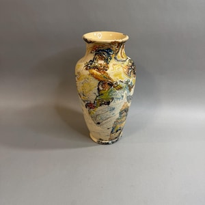 May include: A ceramic vase with a flared rim and a tapered base, featuring a marbled surface pattern in shades of cream, deep blue, mustard yellow, and dark brown. The glaze has a glossy finish with swirling, organic color transitions.