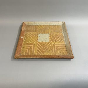 May include: A square ceramic plate featuring a speckled off-white base with a central square and four surrounding rectangular sections, each decorated with embossed chevron patterns in shades of mustard yellow and light brown.