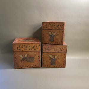 Puede incluir: Tres cajas de madera apiladas, cada una con un diseño de ángel pintado. Las cajas son de color naranja cálido con un borde y un diseño de hojas marrón oscuro. El ángel está representado con alas y un vestido fluido.