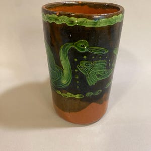 Puede incluir: Vaso de cer&aacute;mica pintado a mano con exterior marr&oacute;n oscuro, base naranja y borde verde. El vaso presenta ilustraciones verdes de un pato y un pez. El dise&ntilde;o recuerda al arte popular.