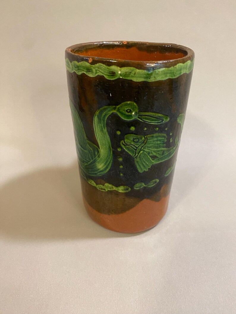 Puede incluir: Vaso de cer&aacute;mica pintado a mano con exterior marr&oacute;n oscuro, base naranja y borde verde. El vaso presenta ilustraciones verdes de un pato y un pez. El dise&ntilde;o recuerda al arte popular.