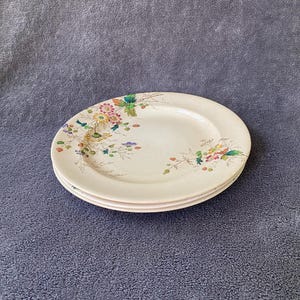 May include: Three off-white oval plates stacked on top of each other. Each plate has a floral design with pink, yellow, and green flowers. The plates are on a gray surface.